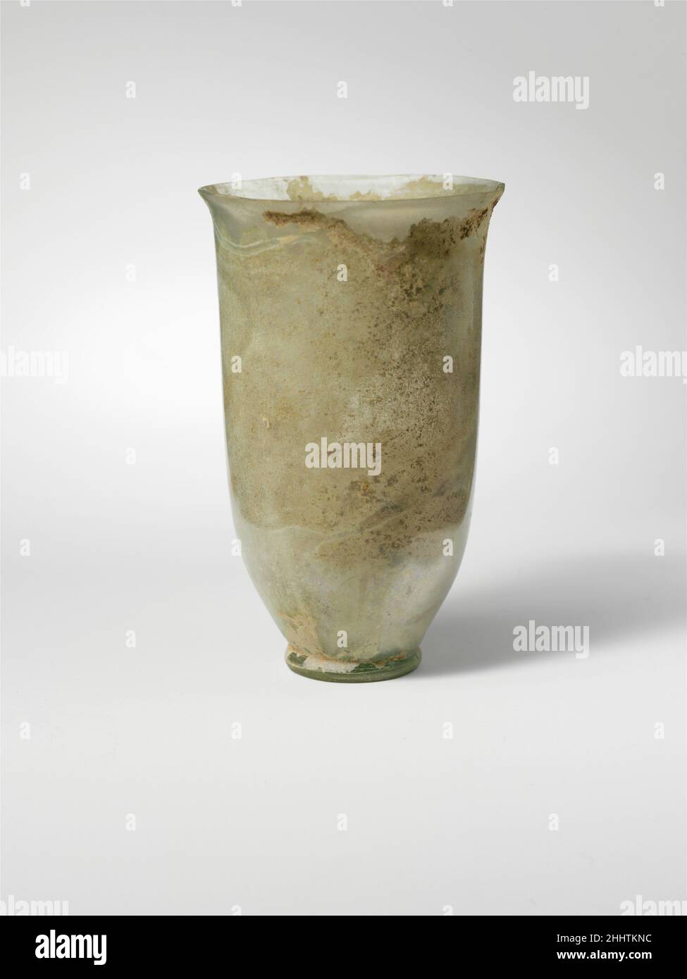 Glass beaker 1st century A.D. Roman Colorless with pale blue green tinge.Knocked-off, uneven rim; slightly bulging collar below rim; narrow, cylindrical body with straight side, then tapering in to folded foot ring; small bottom with low central kick.Intact, except for small chip in rim; some bubbles and blowing striations; iridescence and creamy weathering, with thick soil encrustation around bottom of interior.. Glass beaker  245249 Stock Photo