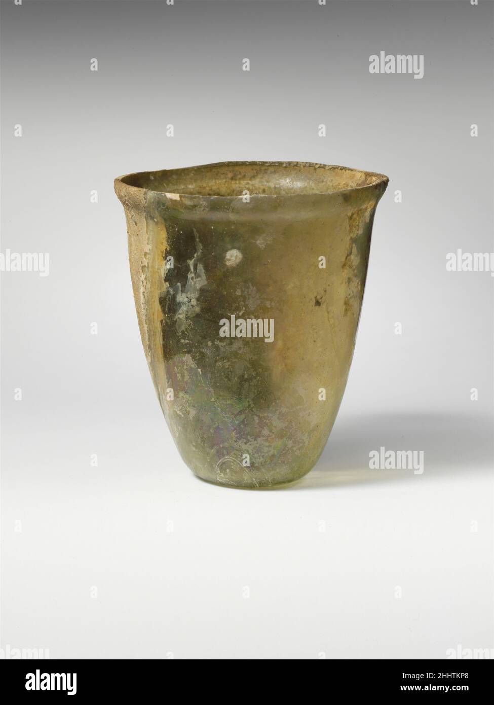 Glass beaker 1st century A.D. Roman Colorless with pale greenish tinge.Knocked-off, uneven rim; slightly bulging collar below rim; convex side tapering downward; small, concave bottom.Intact; pinprick bubbles and blowing striations; brilliant iridescence and faint weathering on one side, but two-thirds of body covered with muddy brown encrustation.. Glass beaker  245246 Stock Photo