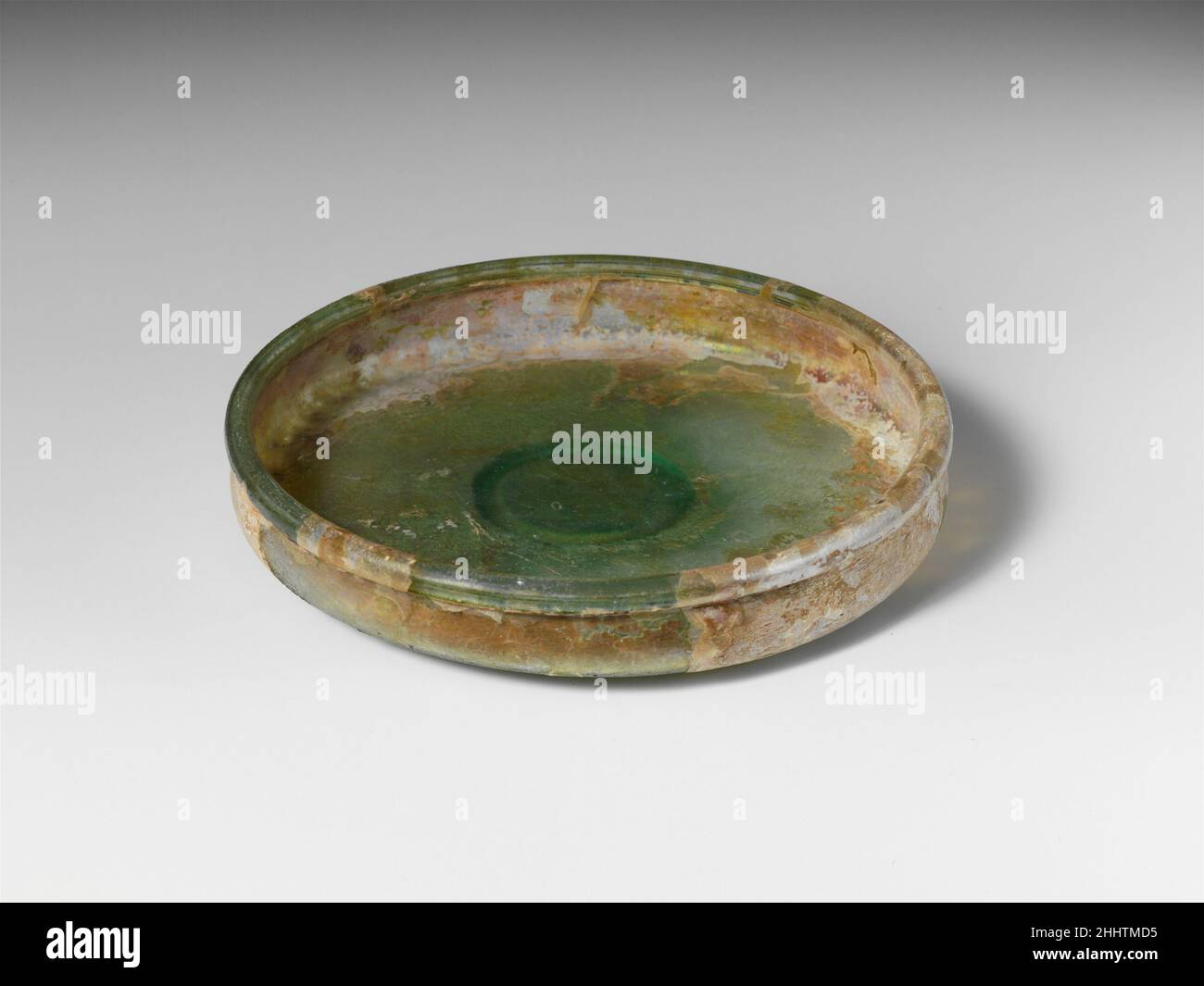 Glass dish 1st century A.D. Roman Translucent blue green.Tubular rim, folded in, round, out, and down; shallow, convex side to body, expanding slightly downwards; broad, almost flat, and slightly pushed-in bottom; small applied solid foot ring, with flat base.Intact; pinprick bubbles; dulling, iridescence, and large patches of creamy weathering.. Glass dish  245295 Stock Photo