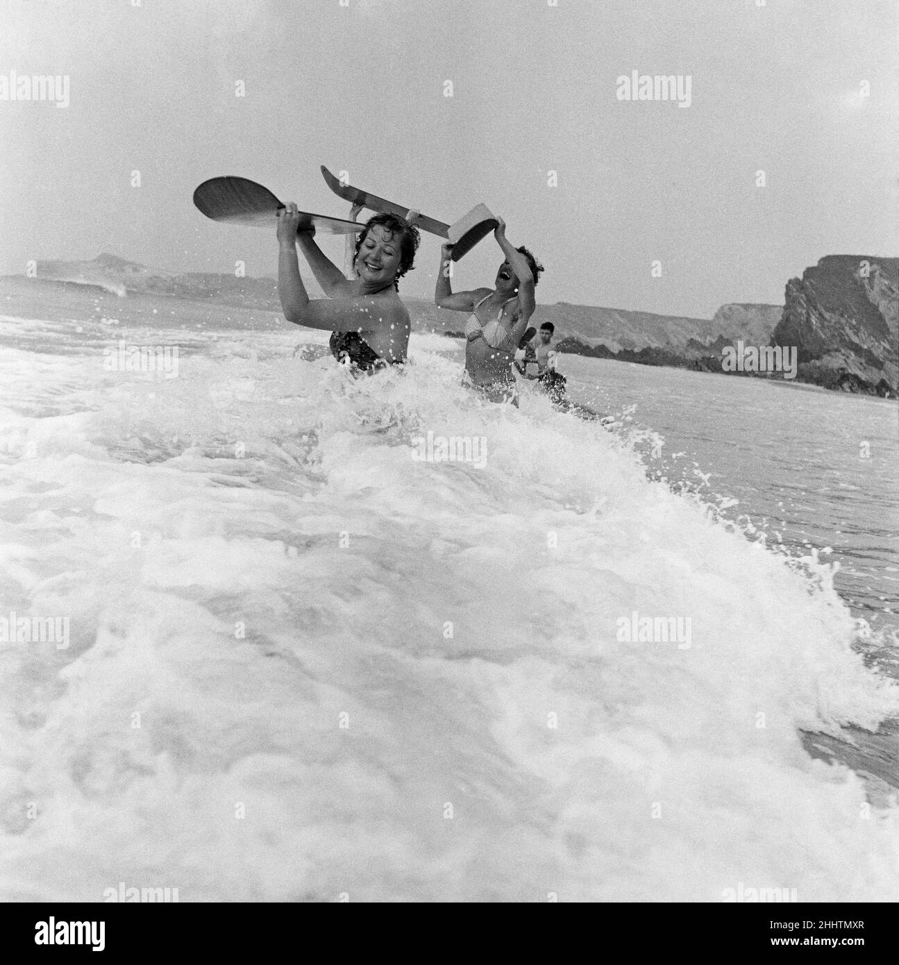 When the foaming waves break with a roar, bathers throw themselves forward onto their surf boards and speed along on the wave crest towards the shore. Pictured, women from Switzerland dash into the foaming surf to reach the breaks in deeper water and be borne inshore on their surf boards. Tolcarne beach, Newquay, Cornwall. 5th June 1954. Stock Photo