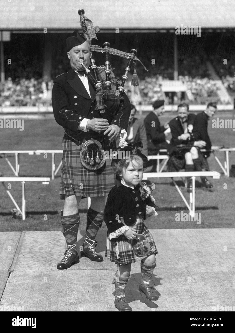 Young Heather Gibb dressed in traditional Scottish tartan costume, entertains the crowd with a dancing routine accompanying a man playing the bagpipes at White City, London. 14th May 1950. Stock Photo
