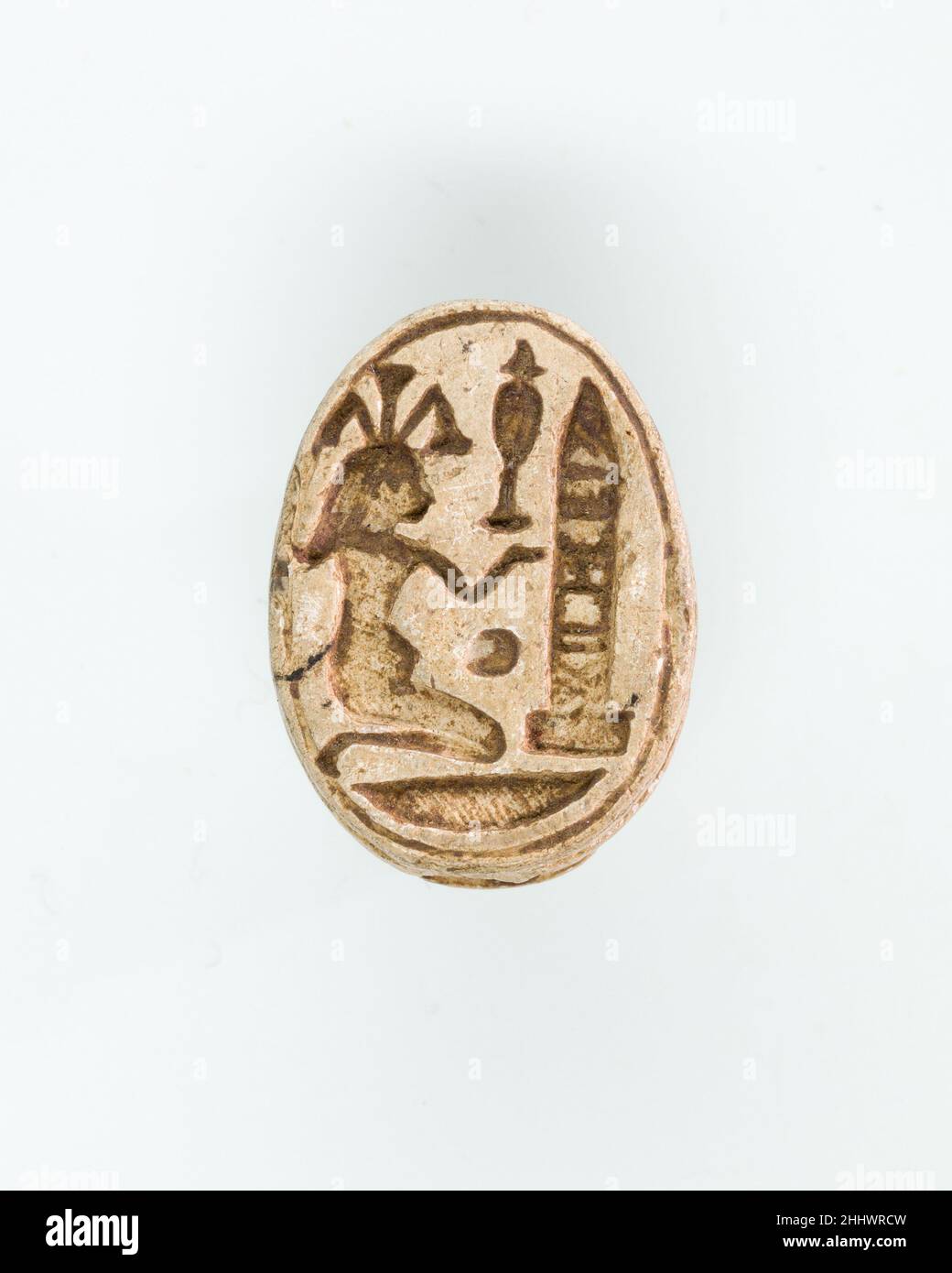 Scarab with kneeling Nile god before obelisk ca. 1295–1070 B.C. New Kingdom or early Third Intermediate Period The underside of the scarab shows the Nile god Hapy, identified by his headdress of papyrus stalks. He is the personification of the annual inundation and therefore also one of the ancient Egyptian fertility gods. He sits on his knees, offering a hes-vase, the hieroglyph of praise, while he faces an obelisk. The obelisk is one of the symbols of the sun god and can thus stand for Amun. Within the monument, however, one can also discern small hieroglyphs forming the throne name of phara Stock Photo