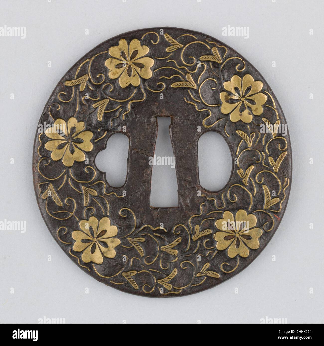 Sword guard (Tsuba) With Cherry Blossom Motif (桜図与四郎象嵌鐔) 17th century Japanese This Yoshirō style iron tsuba shows on both sides a brass inlay (shinchū-zōgan) of cherry blossoms embedded into arabesques. The arrangement of the design suggests that the two openings for scabbard accessories (hitsu-ana) are orignal and were not added later as it is so often the case. In addition, there is almost no loss of inlay and so this tsuba is in excellent condition.. Sword guard (Tsuba) With Cherry Blossom Motif (桜図与四郎象嵌鐔)  29979 Stock Photo