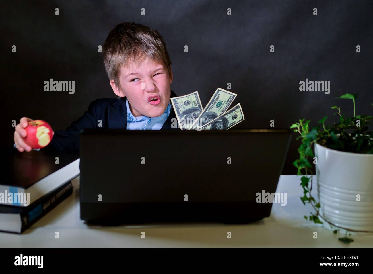 A happy boy in a school suit with dollar money at the computer, copy space on a dark studio background Stock Photo