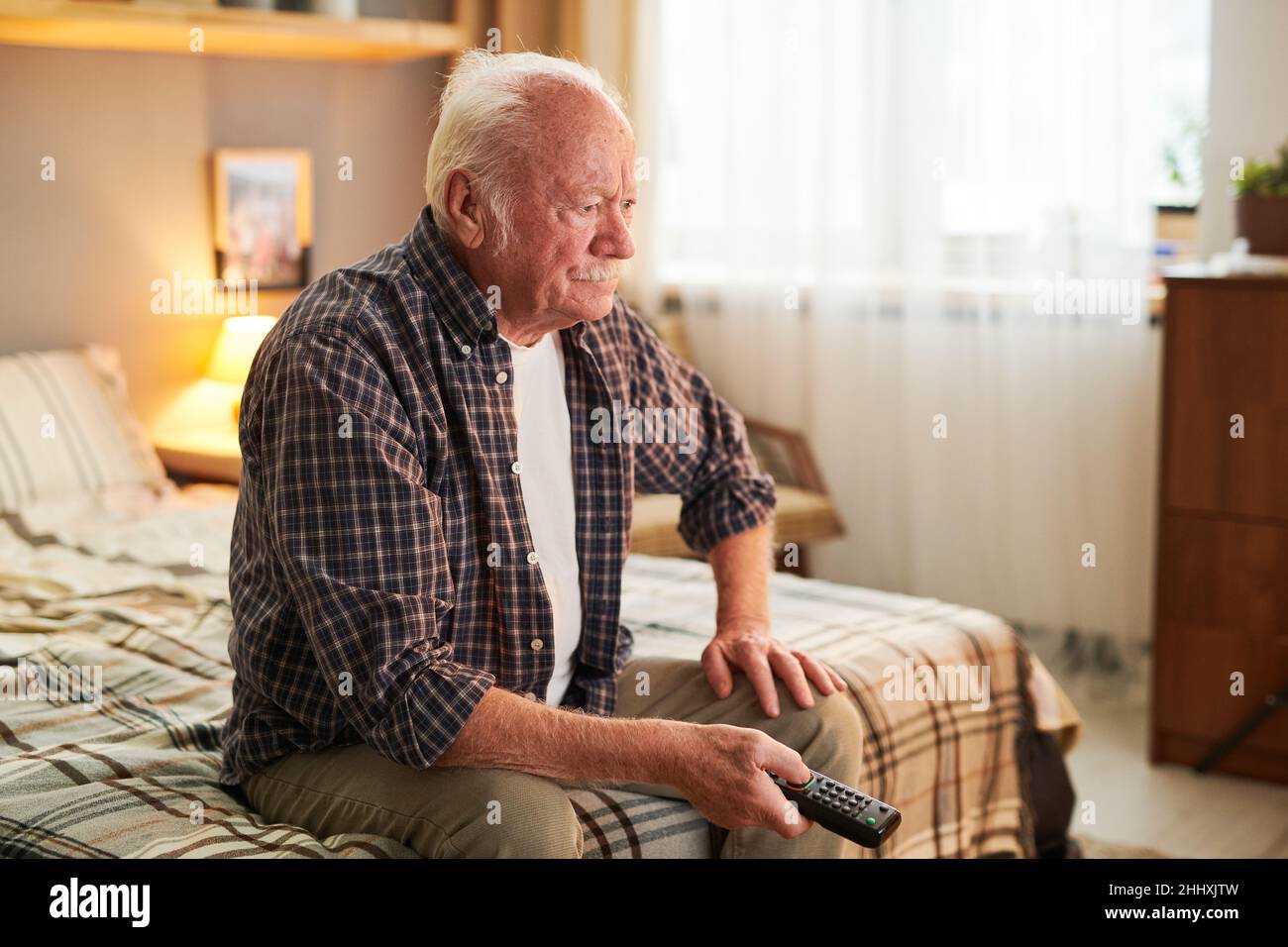 Old man with remote control choosing channel while sitting on double bed in his bedroom and watching tv Stock Photo