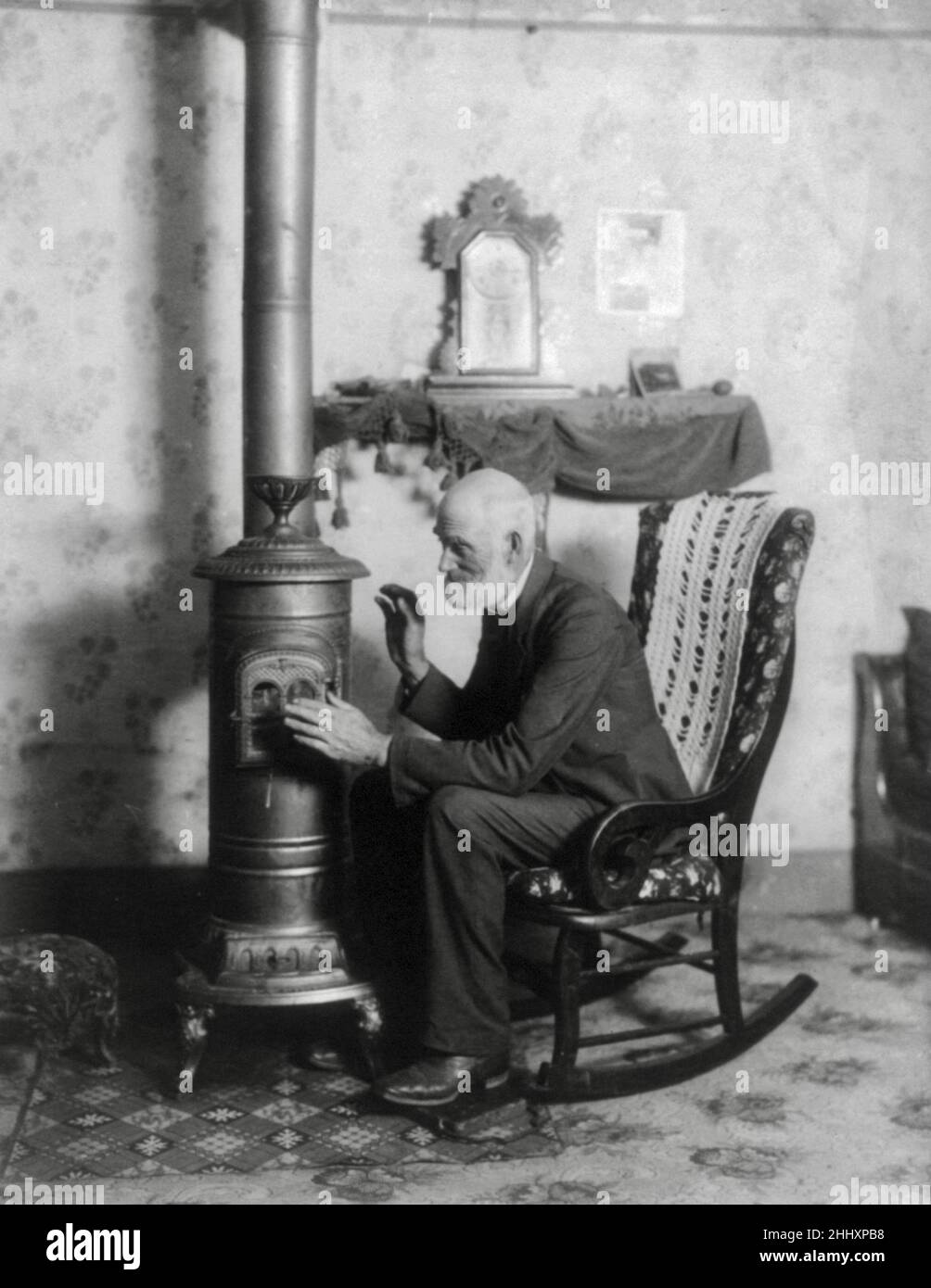 Elderly gentleman keeping warm close to stove. People still struggling to keep warm 100+ years later. Photograph from 1900. Stock Photo