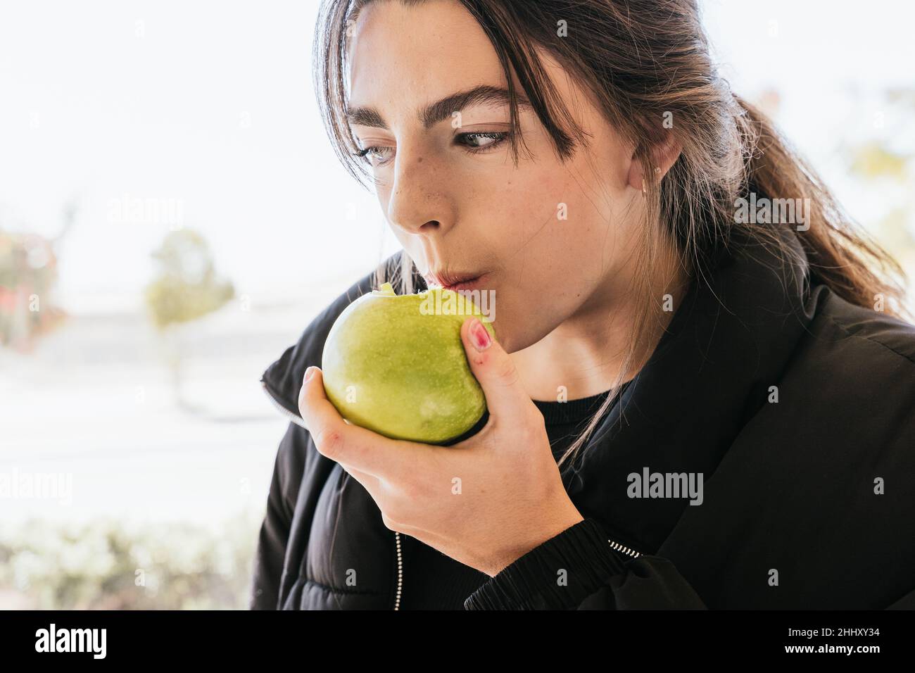 Young female in outerwear biting ripe apple while having healthy lunch on blurred background on sunny day Stock Photo