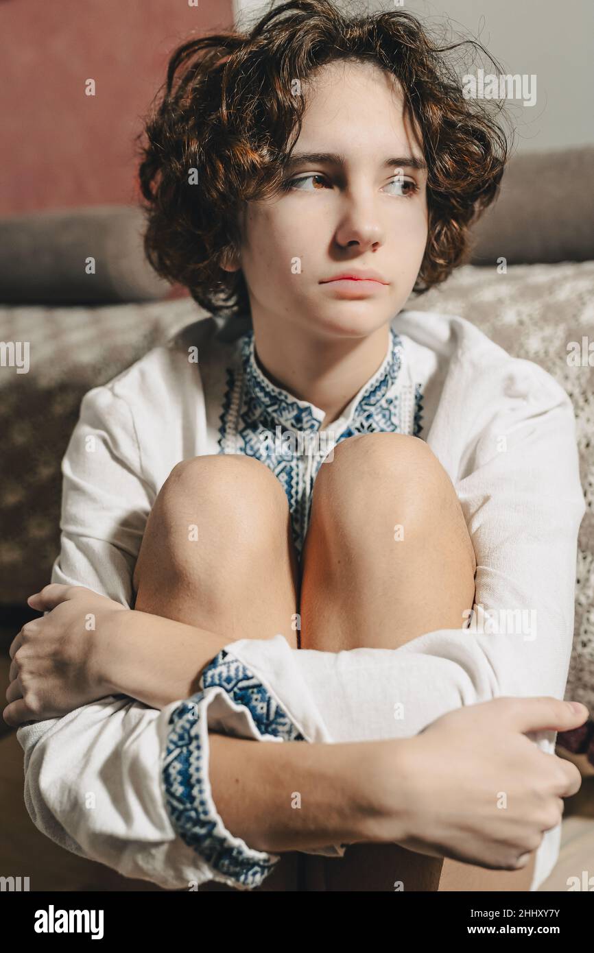 sad young woman with brown curly hair tied in bun, wearing white ethnic embroidered shirt, sits in depressed pose, bending legs, clasped hands. concep Stock Photo