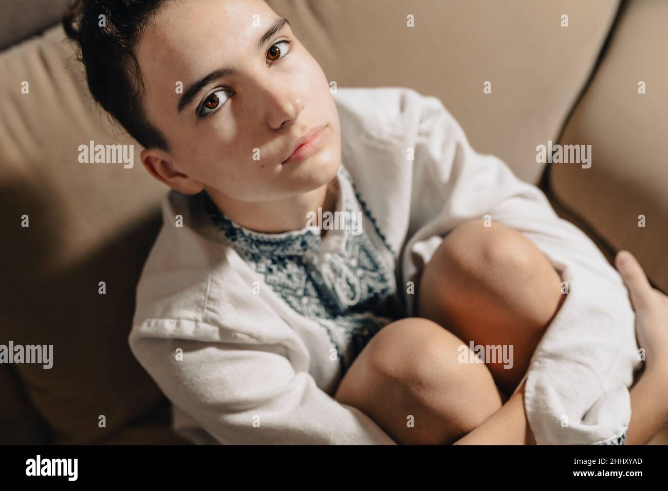 teenage girl with brown hair gathered in bun, with shaved temples, in white ethnic embroidered shirt, sad emotion on face, sits cross-legged and huggi Stock Photo