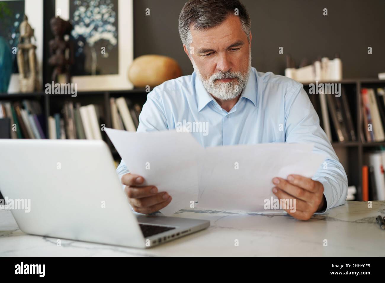 Serious mature male employee sits at the table in modern office, works with documents. Concentrated business man is using laptop, browsing internet, s Stock Photo
