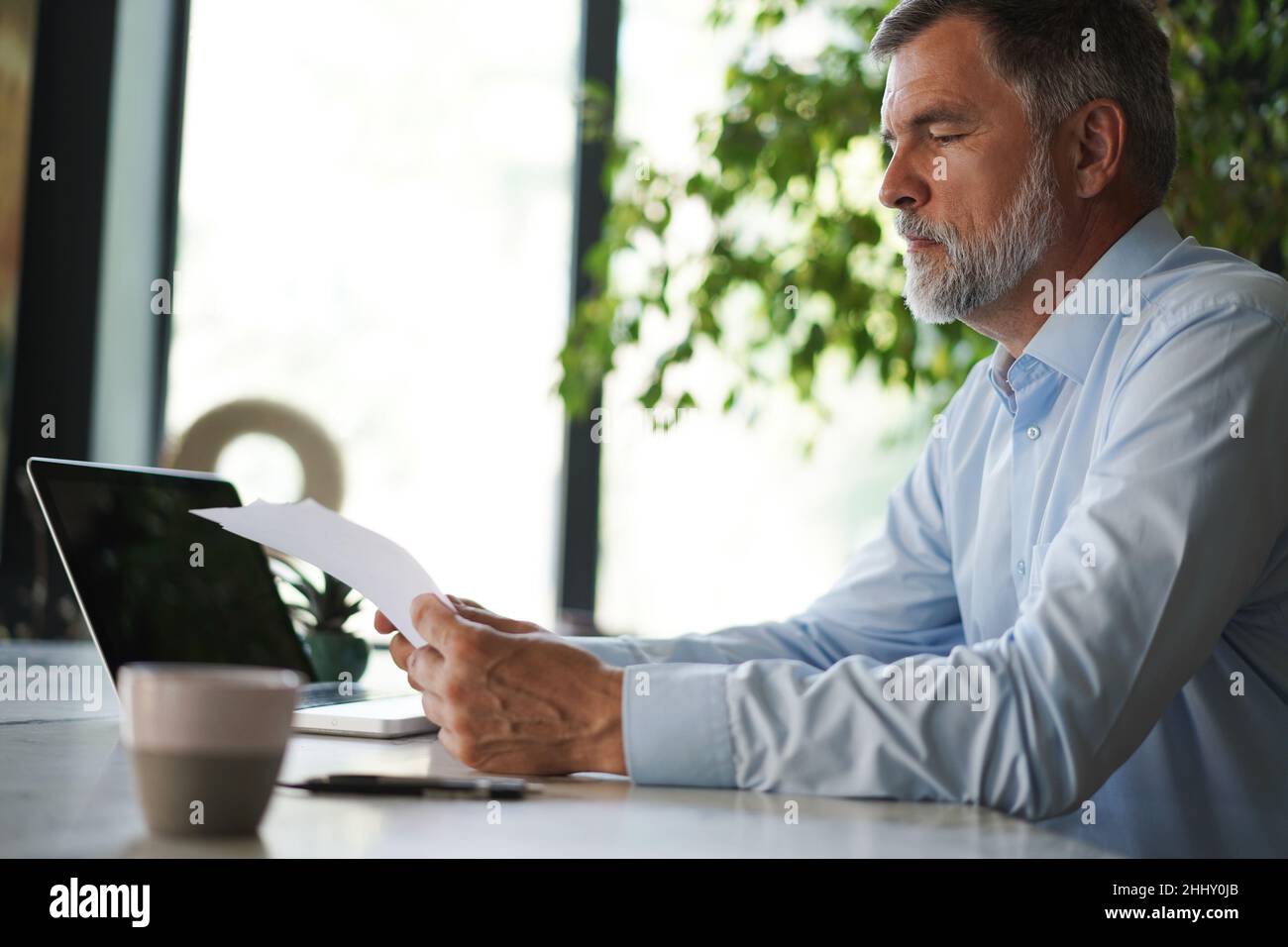 Serious mature male employee sits at the table in modern office, works with documents. Concentrated business man is using laptop, browsing internet, s Stock Photo
