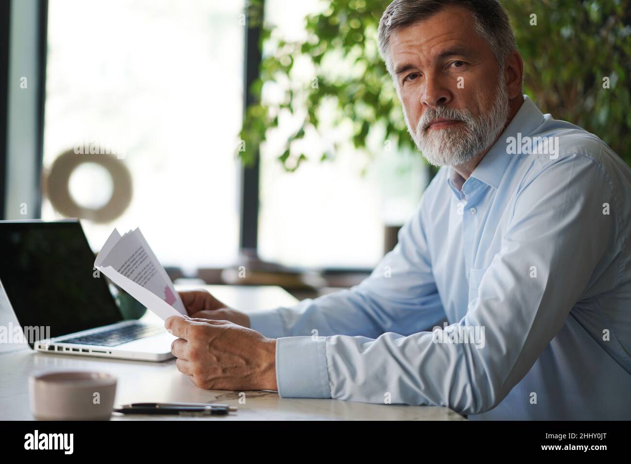 Serious mature male employee sits at the table in modern office, works with documents. Concentrated business man is using laptop, browsing internet, s Stock Photo
