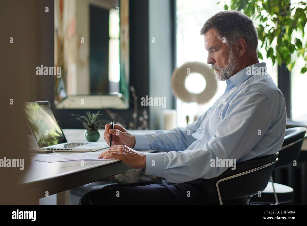 Serious mature male employee sits at the table in modern office, works with documents. Concentrated business man is using laptop, browsing internet, s Stock Photo