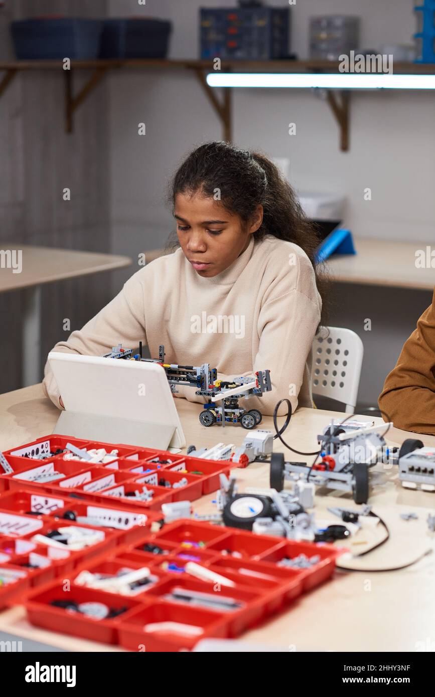 African teenage girl sitting at the table using digital tablet to project robot from constructor at science lesson Stock Photo