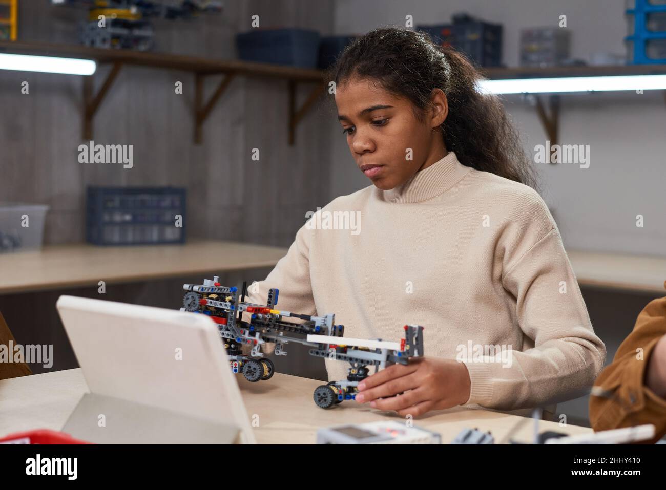 African girl sitting at the table in front of the tablet pc and making robot from constructor at science school Stock Photo