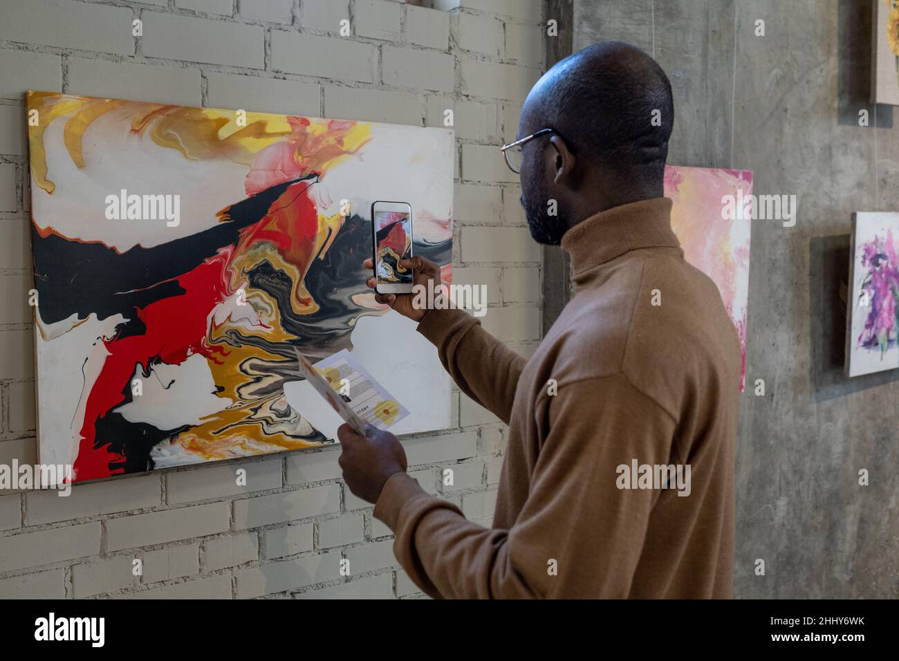 Rear view of African man using his mobile phone to make photo of artwork on the wall during exhibition Stock Photo