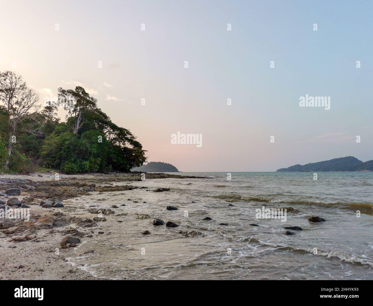 Seascape with boulders and sea waves lash line impact rock on a summer day. Beautiful tropical beach with sunset sky and small island view at horizon. Stock Photo