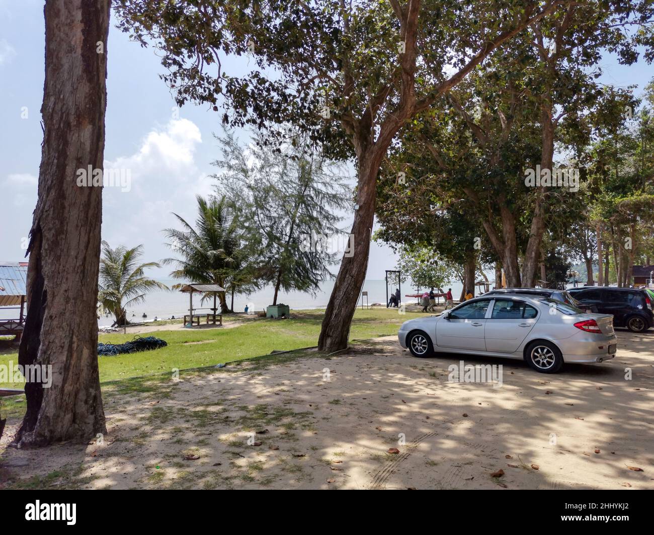 A car parked at sandy beach with grass meadow, blue sky, sea wave, palm tree in the background. People rest on tropical beach. Beautiful coast. Countr Stock Photo