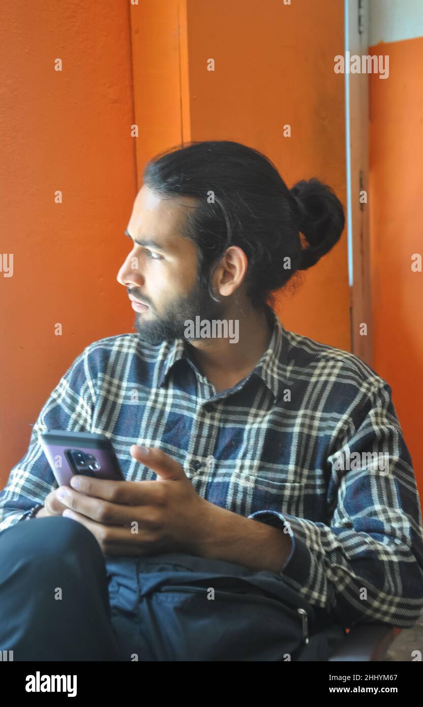 Selective focus of a good looking bearded man with hair bun, looking sideways during using his phone while sitting indoors Stock Photo