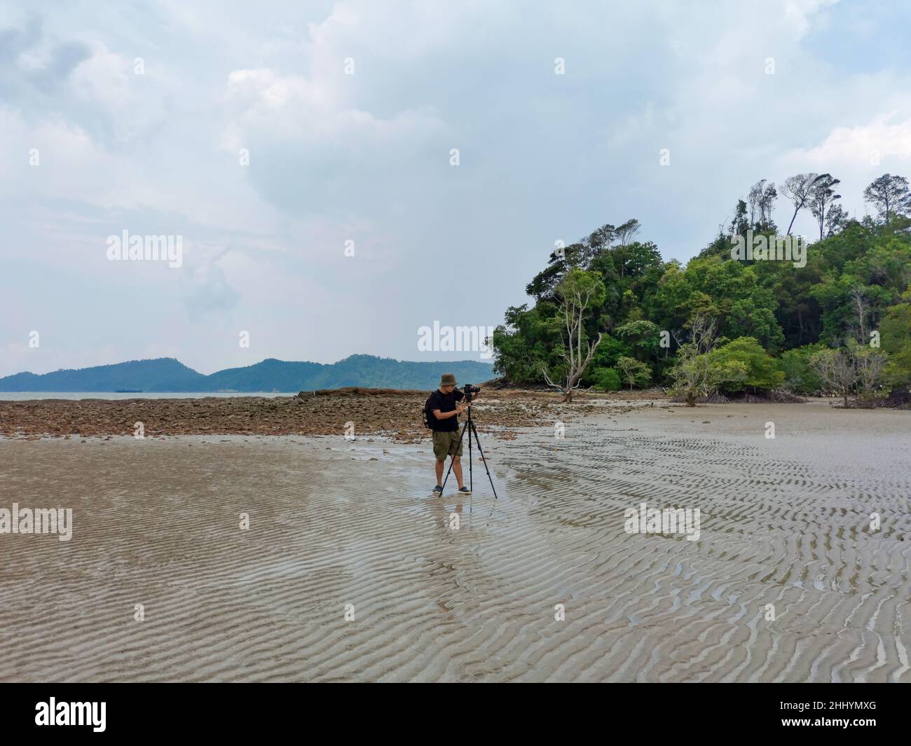 Nature photographer in the action. Alone traveller shoots the seascape with mangrove rainforest background. Backpacker take photo on low tide sandy be Stock Photo