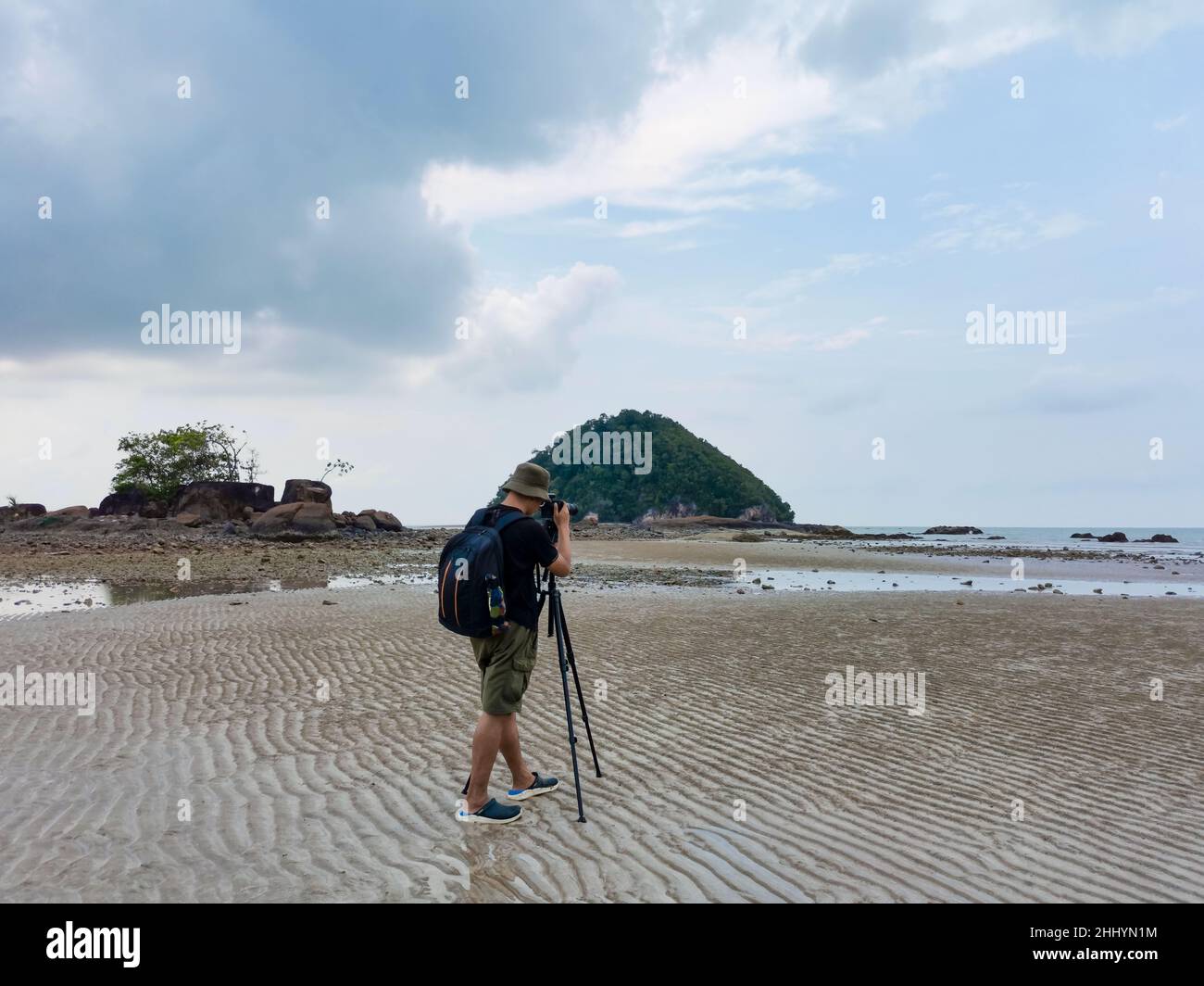 Nature photographer in the action. Alone traveller shoots the seascape with small island background. Backpacker take photo on low tide sandy beach. En Stock Photo