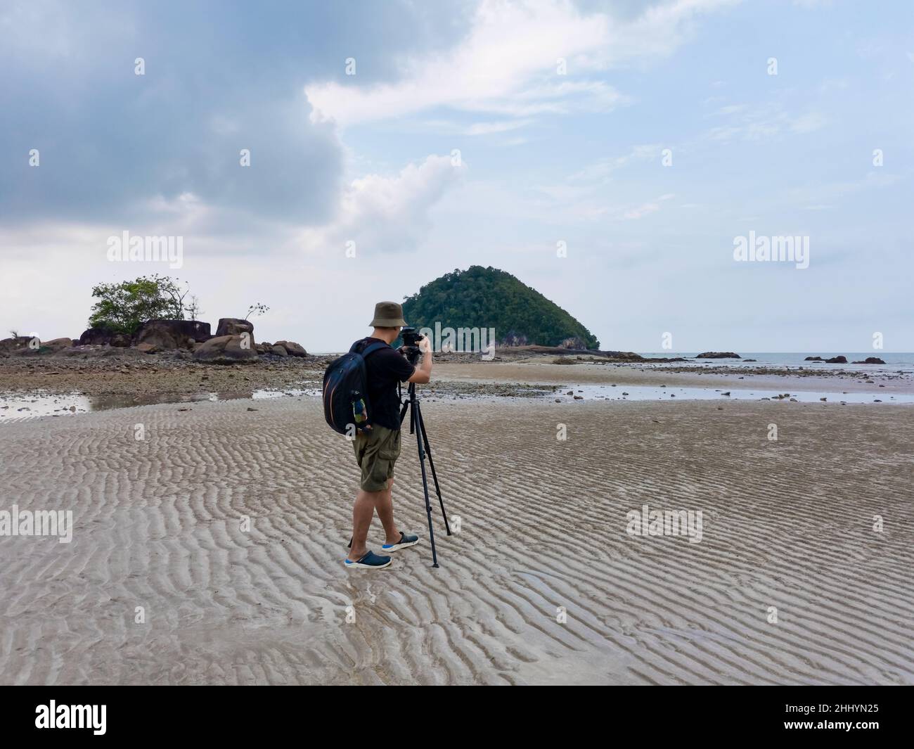 Nature photographer in the action. Alone traveller shoots the seascape with small island background. Backpacker take photo on low tide sandy beach. En Stock Photo