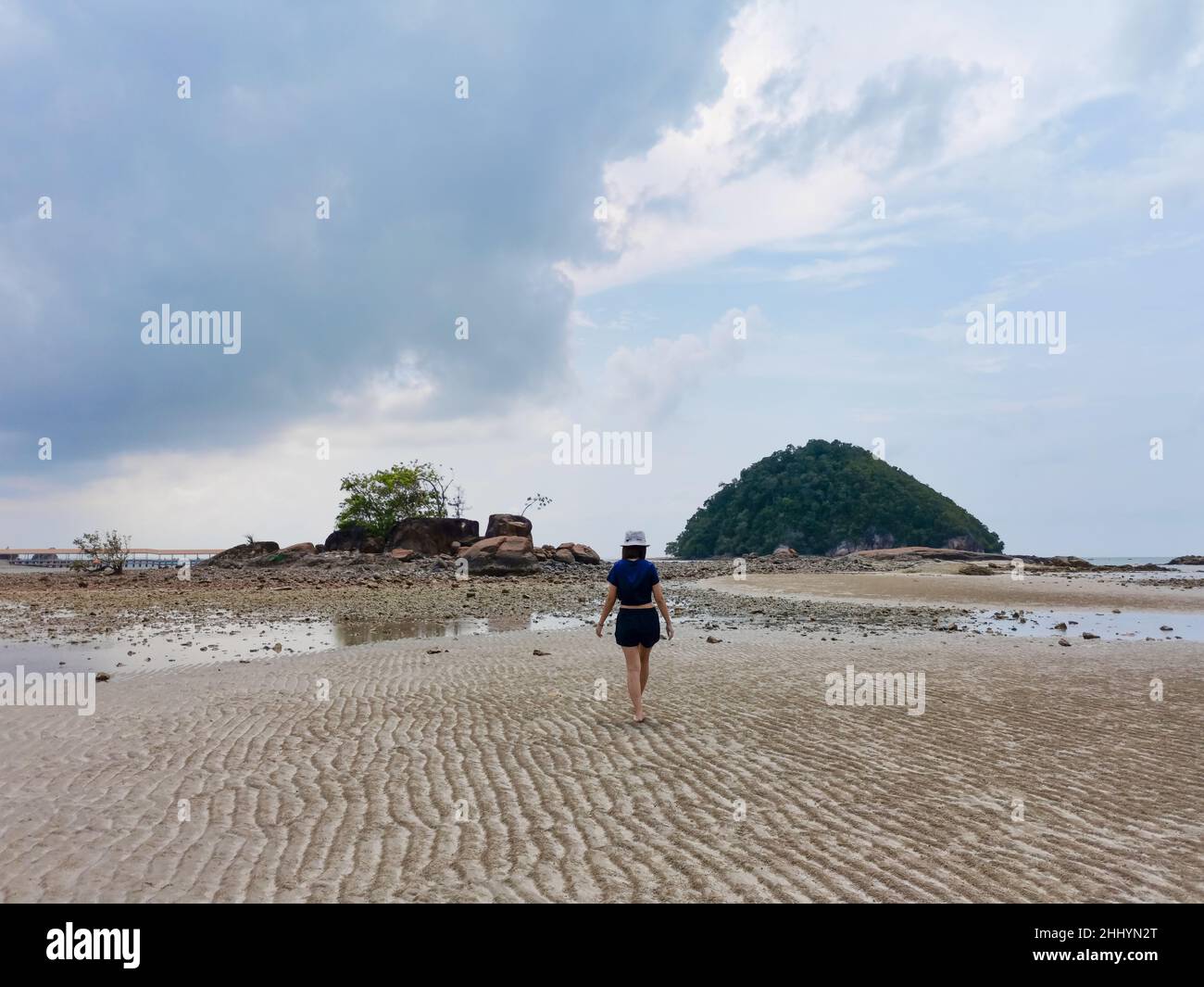 Traveller asian woman walking in a calm and tranquil sandy beach at low tide with small island and rock mountain and gravel and cloudy sky background, Stock Photo