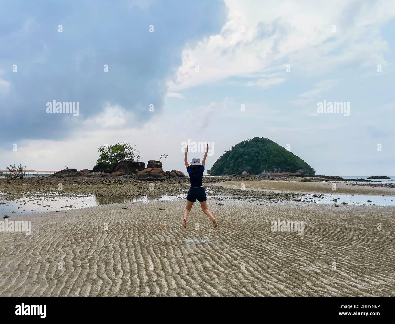 Happy girl traveler jumping on beach at low tide with small island and rock mountain and cloudy sky background. Freedom and happiness concept. Travel Stock Photo