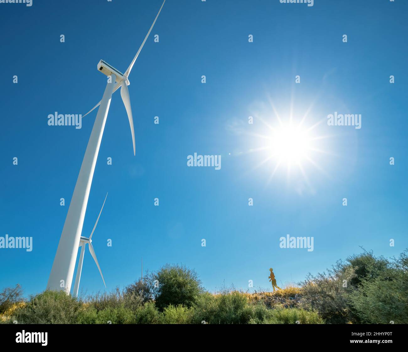 Huge wind turbine and tiny human silhouette on top of a hill against blue sky and shining sun, with lens flare Stock Photo