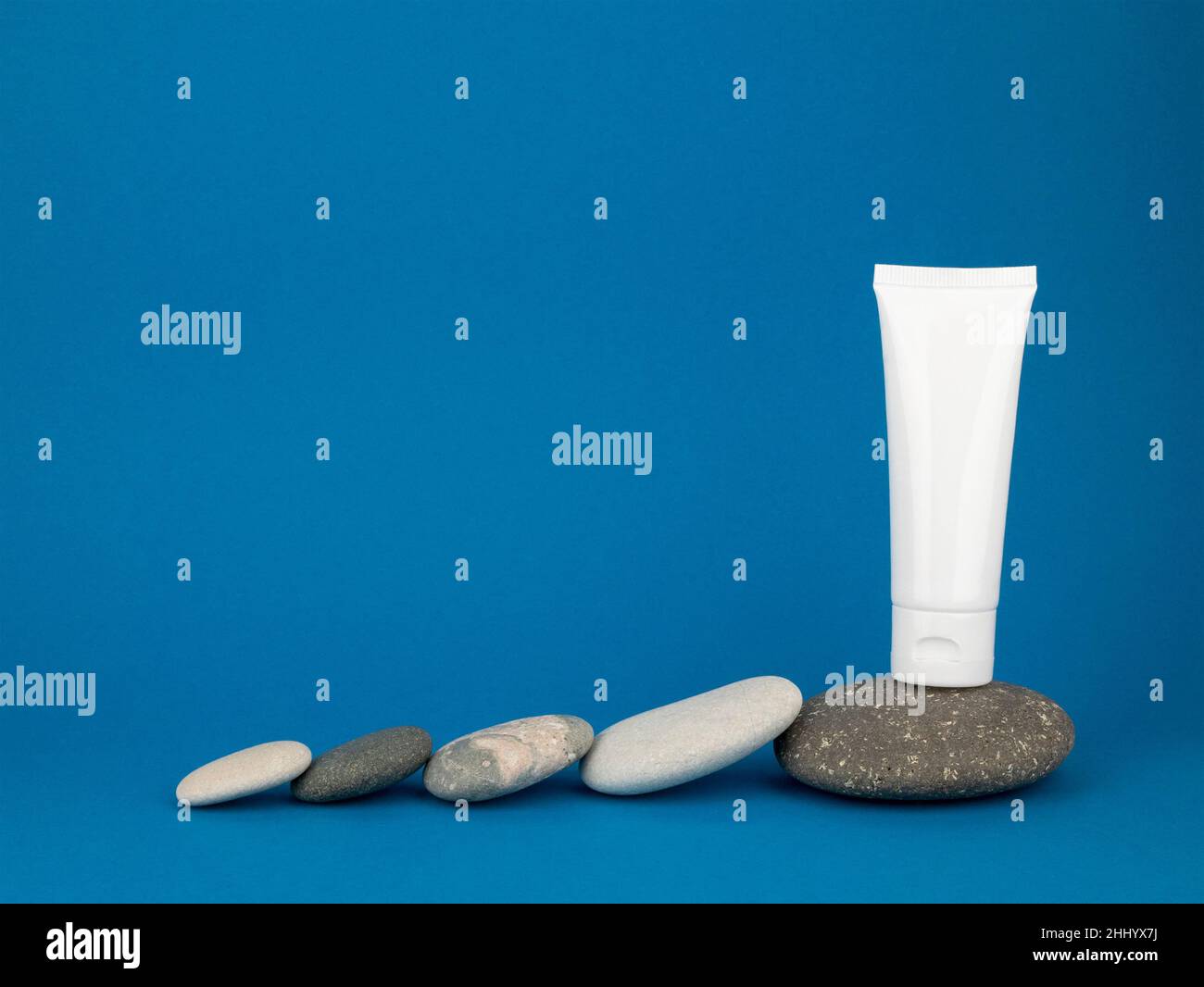 White tube and rounded pebbles on blue background, copy space. Mockup plastic tube for sunscreen or moisturizing cream Stock Photo