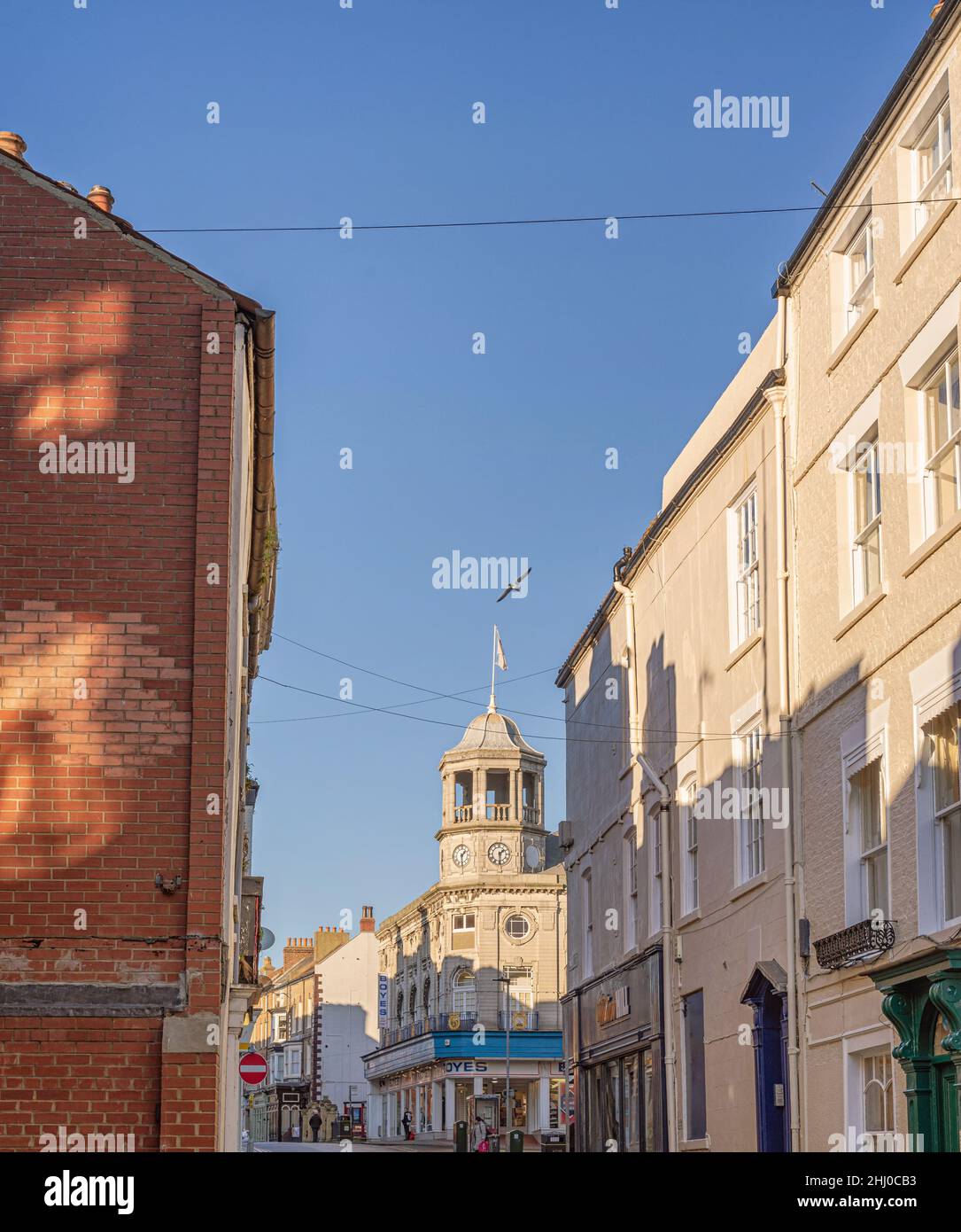 View along a narrow street to a building with a tower and dome.  The sunlight throws shadows onto a wall and a blue sky is above. Stock Photo