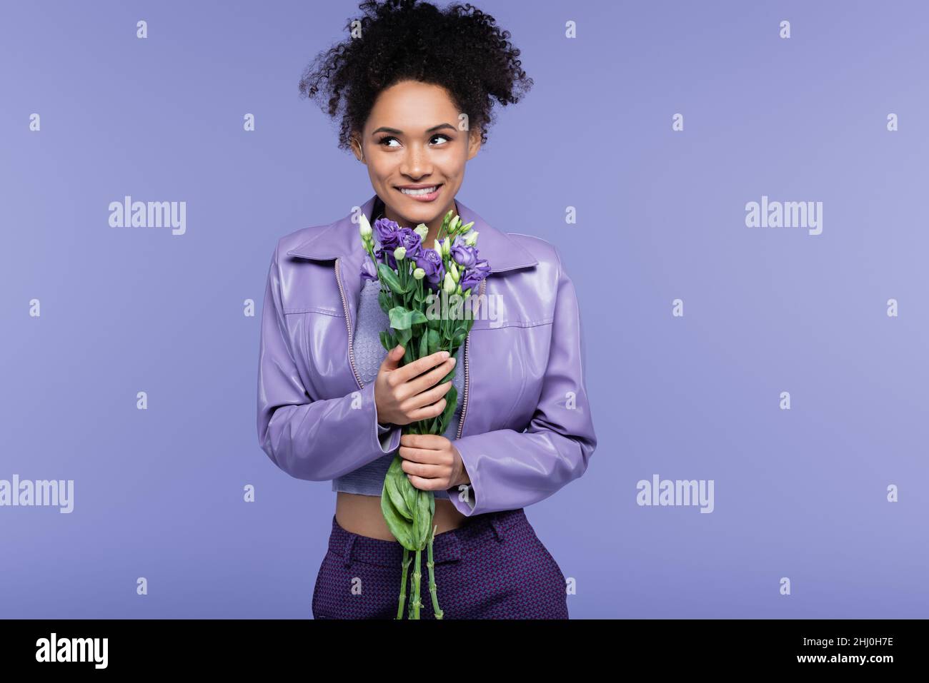 happy young african american woman biting lip and holding bouquet of flowers isolated on purple Stock Photo