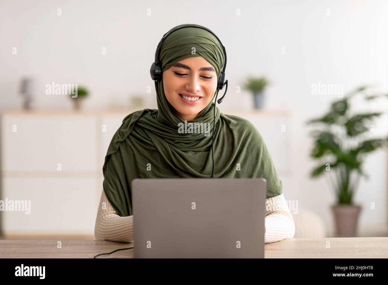 Millennial Arab lady in hijab using laptop at home, wearing headphones ...