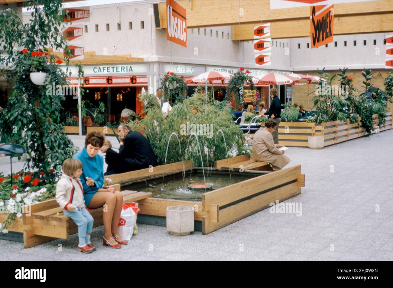 Undercover shopping at the Tarup Center on the western outskirts of Odense, Funen, Denmark in 1970. Red and white signs indicate that it is summer sale (sommer udsalg) time. The use of timber in the centre’s design is evident. The centre opened in 1968 with 21 stores and was the first covered shopping centre on Funen. It is now much expanded with over double the number of stores at the site and it has been extensively remodelled. Odense is the main city of the island of Funen. This image is from an amateur 35mm colour transparency – a vintage 1970s photograph. Stock Photo