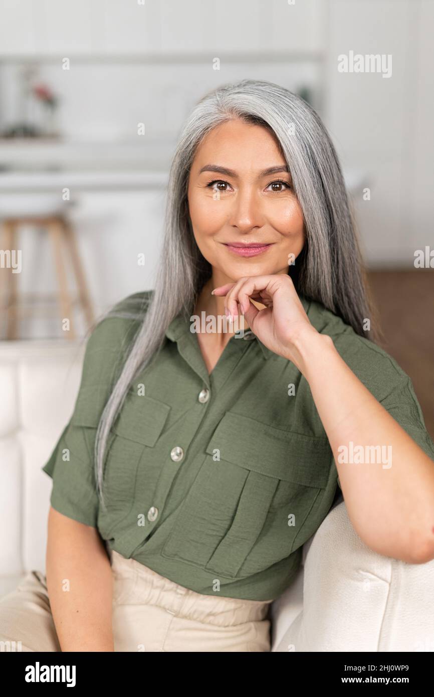 Headshot of smiling good-looking senior Asian female looking at camera. Cheerful carefree grey-haired woman resting at home, vertical shot Stock Photo
