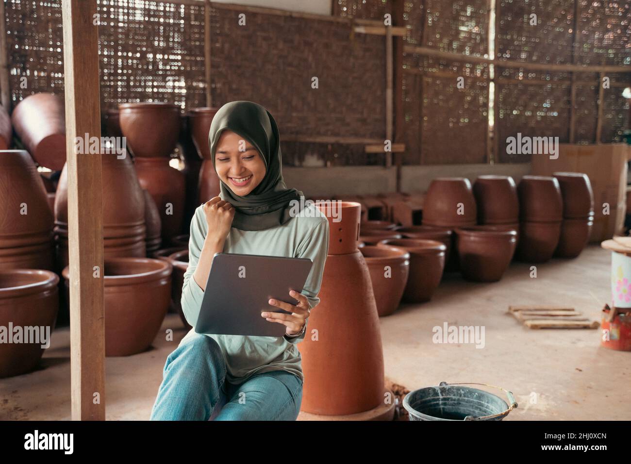 veiled asian woman smiling using tablet while sitting between pottery Stock Photo