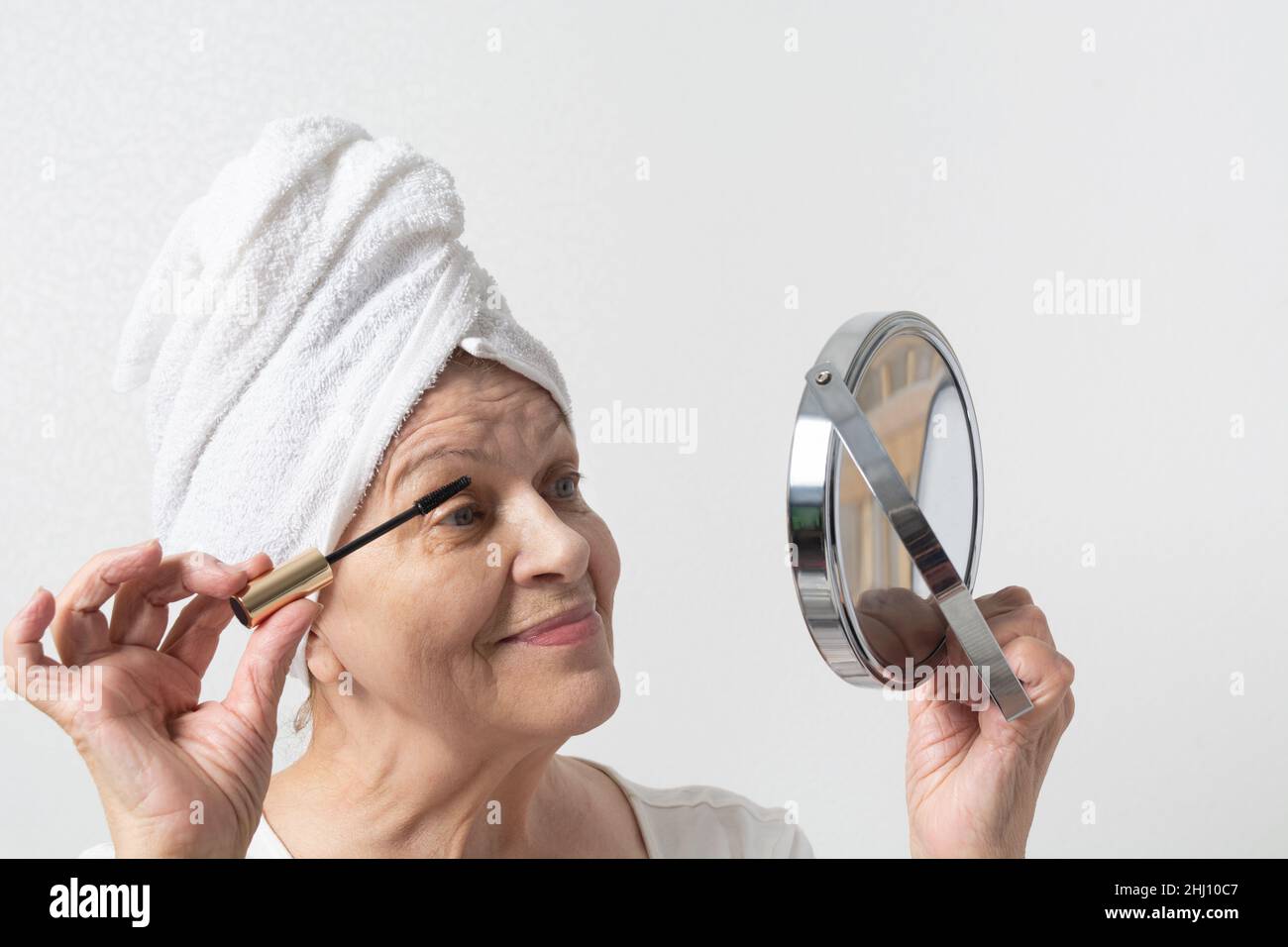 Happy elderly lady with towel on head coloring eyelashes and looking at mirror Stock Photo