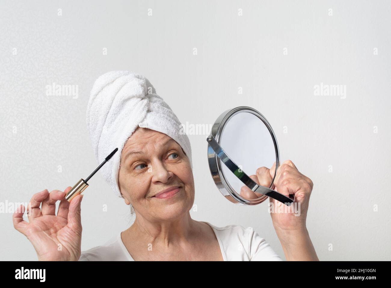 Happy elderly lady with towel on head coloring eyelashes and looking at mirror Stock Photo