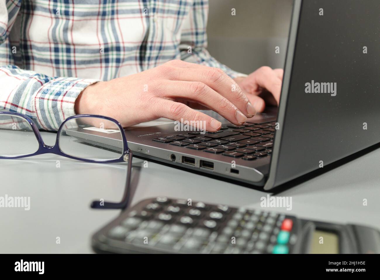 Office worker in checked shirt working at laptop with glasses and calculator on desk Stock Photo