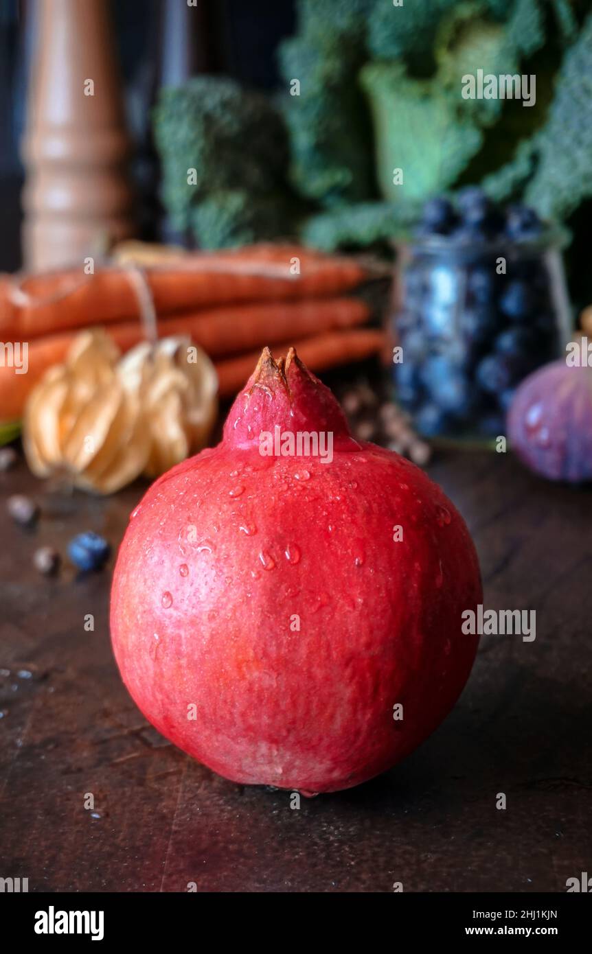 A whole ripe pomegranate with water drops on the background of autumn vegetables and berries Stock Photo