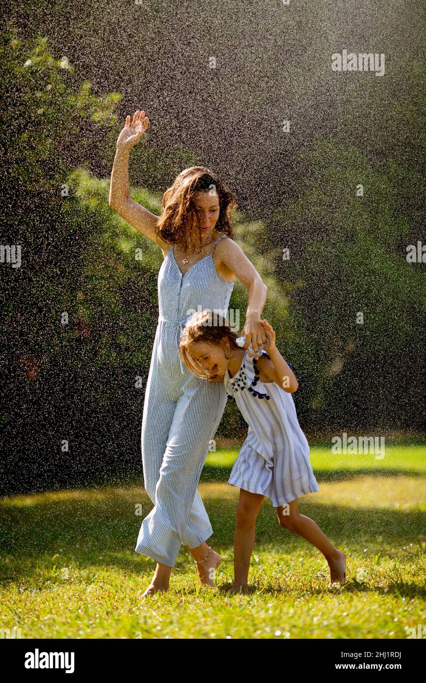 Cute little girl having fun with water under irrigation sprinkler with her mother Stock Photo