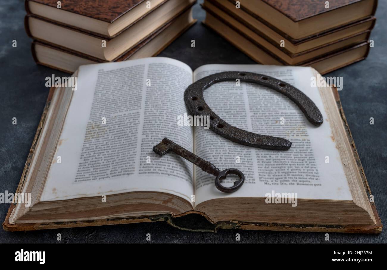 An old key and an old horseshoe rest on the pages of an open ancient book, with other books in the background Stock Photo