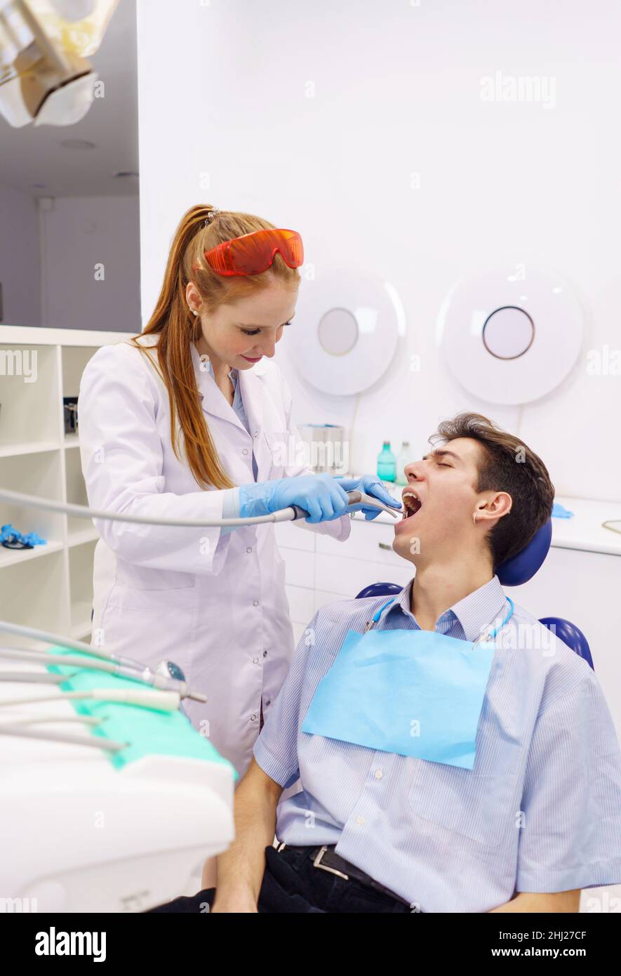 Redhead woman in medical uniform using dental drill to heal teeth of male patient in light dentist office of clinic Stock Photo
