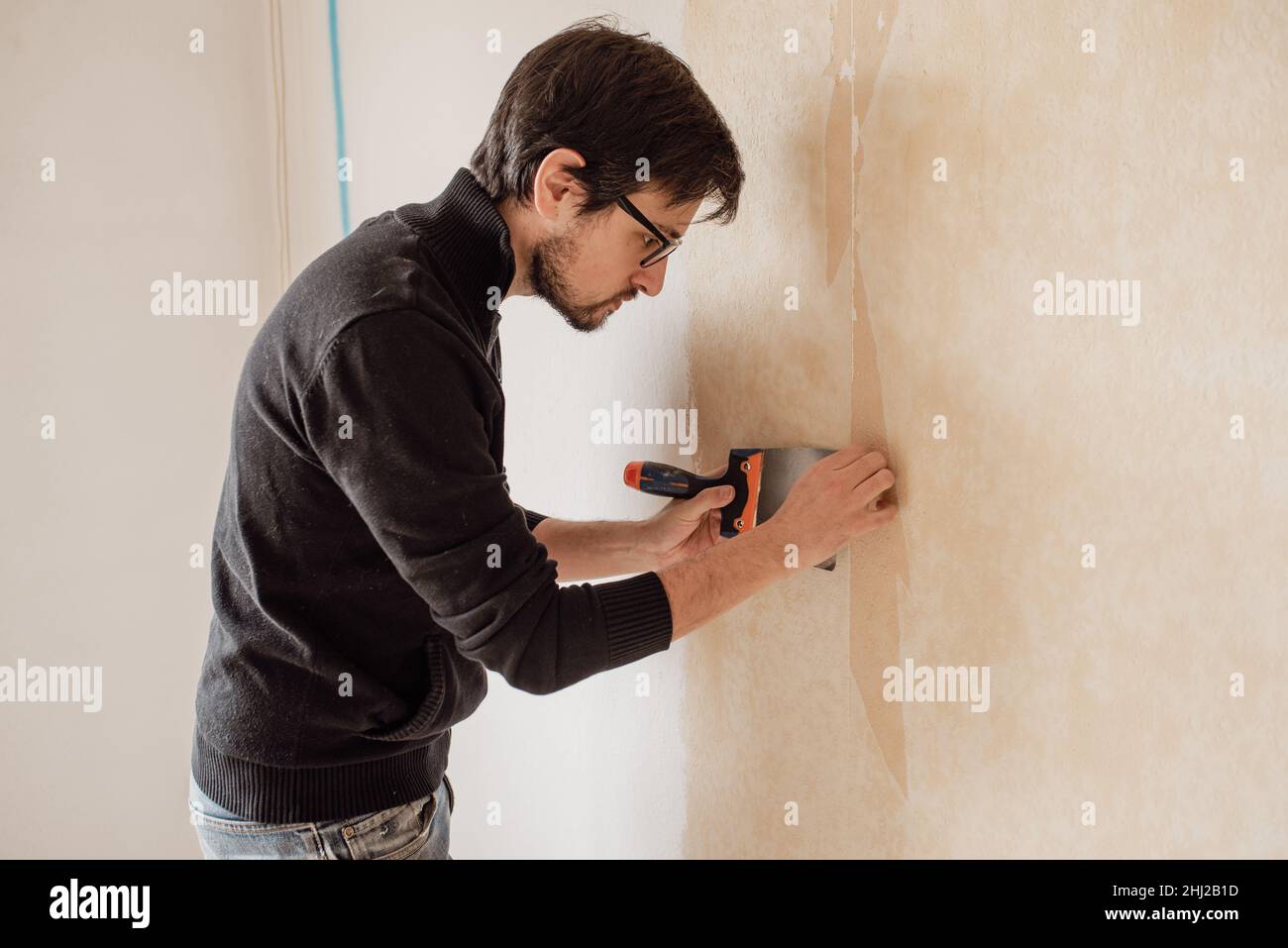 Man hand tearing off old wallpaper with scraper from wall. Home renovations. Stock Photo
