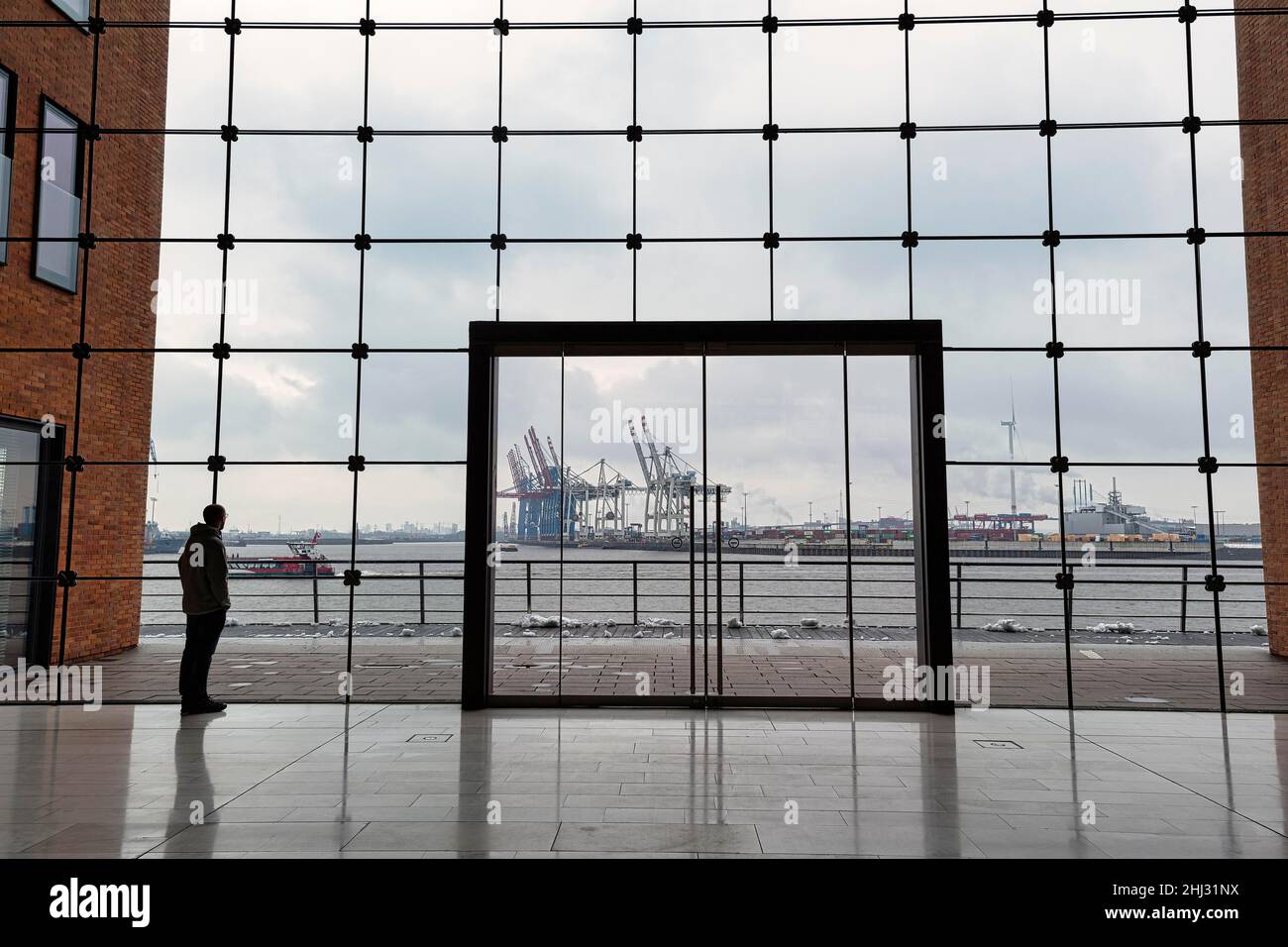 Young man standing in front of modern glass facade with large door and looking at the harbour, dreary winter weather, backlight, Holzhafen Ost Stock Photo