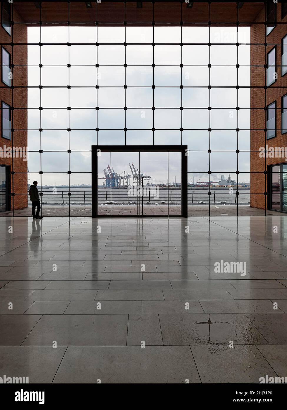 Young man standing in front of modern glass facade with large door and looking at the harbour, dreary weather, backlight, Holzhafen Ost office Stock Photo