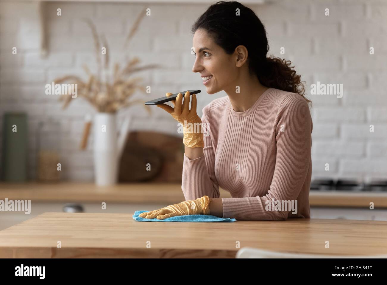 Latin housewife holds smartphone talks on speakerphone while cleaning kitchen Stock Photo