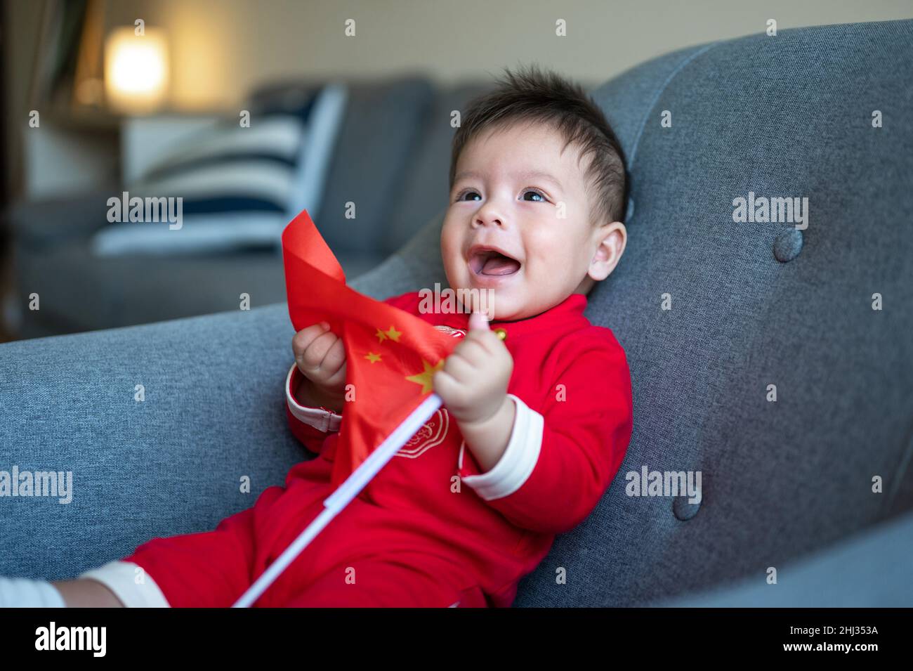 Asian baby boy holding Chinese flag sitting on a sofa while wearing traditional Chinese red clothes for Lunar New Year at home Stock Photo