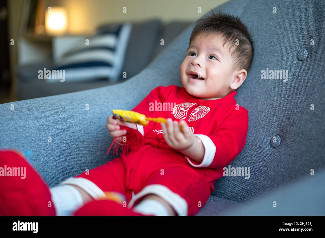 Baby boy holding a Chinese knot for good luck while sitting on a sofa wearing traditional Chinese red clothes for Lunar New Year Stock Photo