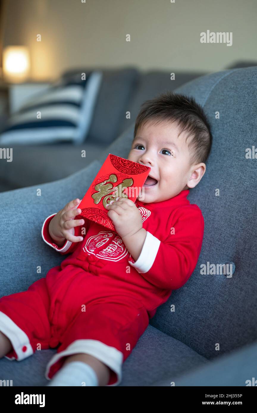 Asian baby boy holding a red pocket envelope for Lunar New Year sitting on a sofa wearing traditional Chinese red clothes at home Stock Photo